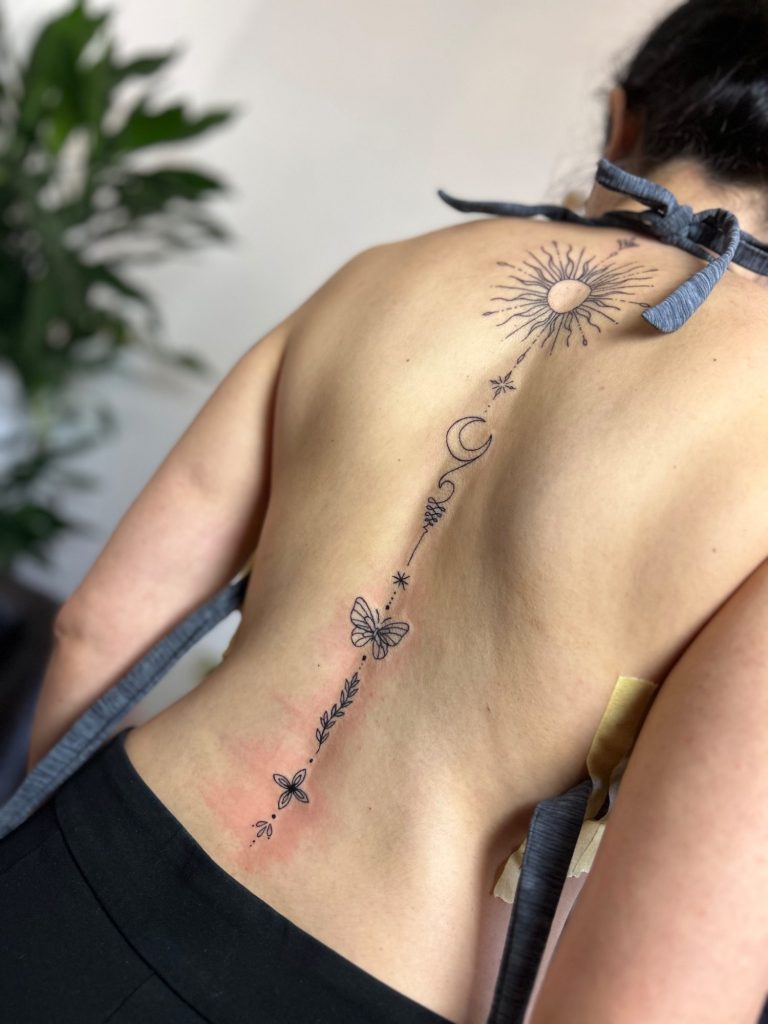 Fineline Rücken Tattoo mit Sonne, Mond, Sternen und botanischen Elementen entlang der Wirbelsäule