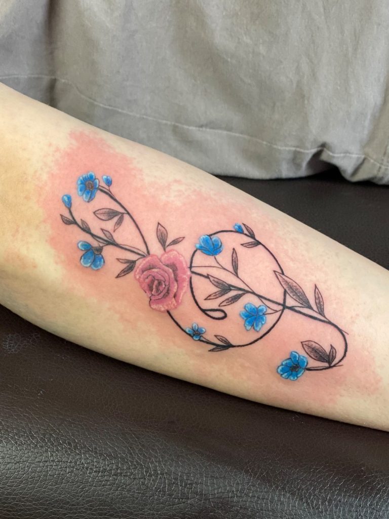 Florales Fineline Tattoo mit Notenschlüssel, Rose und blauen Blüten am Unterarm