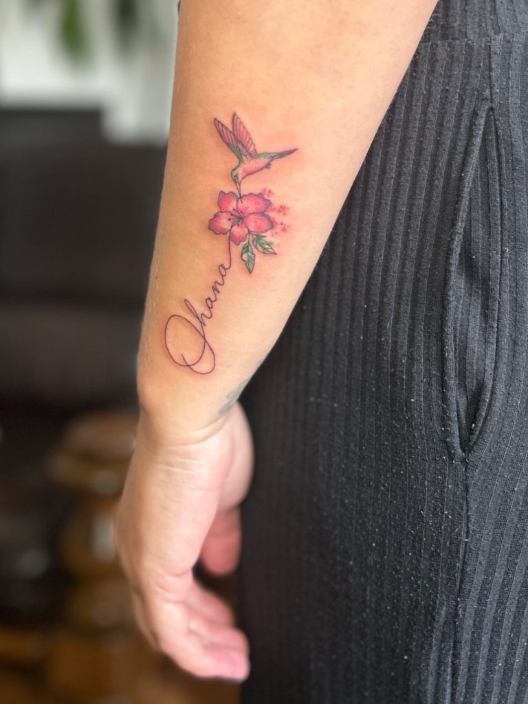 Kleines Aquarell Kolibri Tattoo mit roten Blüten und feinem Schriftzug am Unterarm