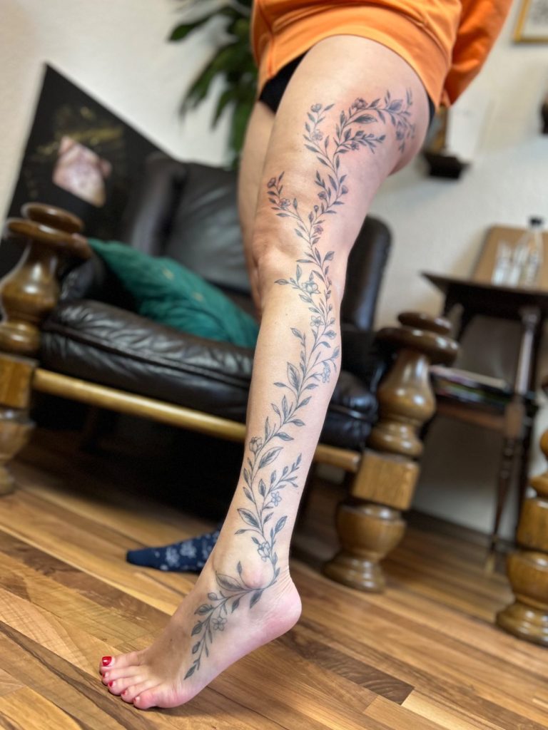 Botanisches Fineline Tattoo mit feinen Blüten und Blättern entlang des Beins